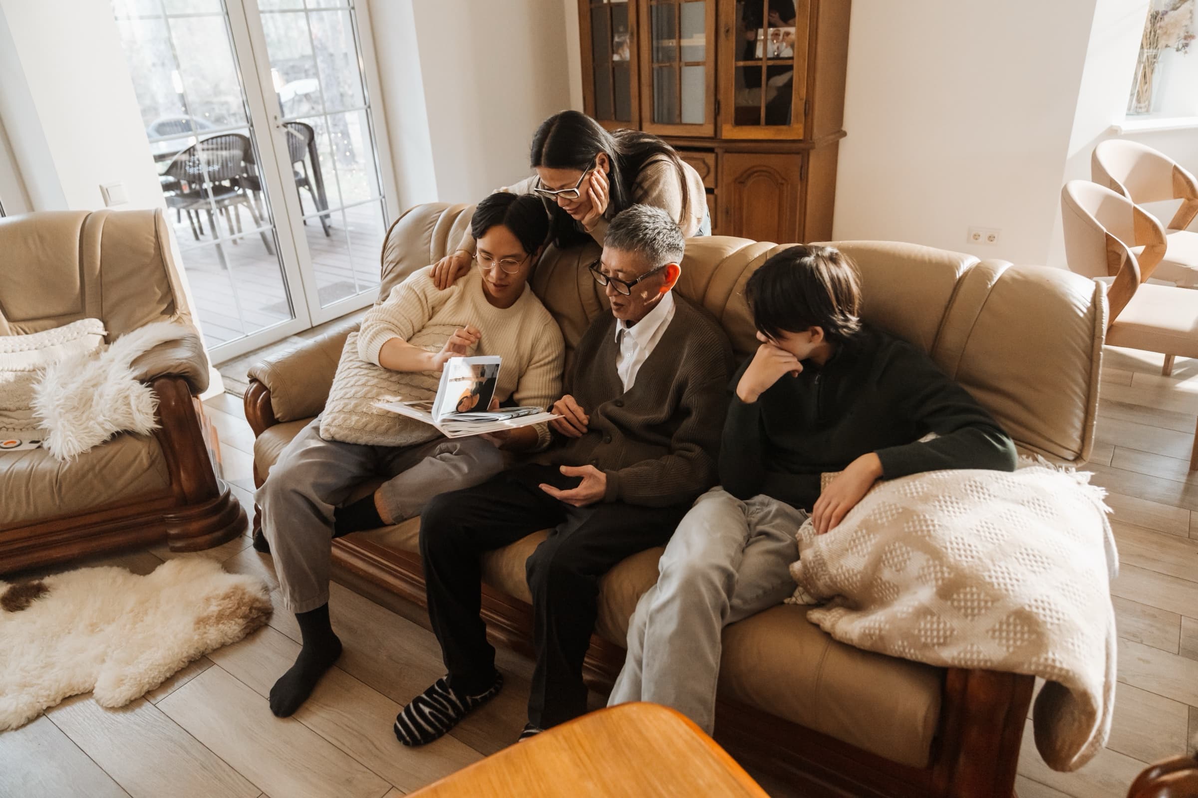 A multi-generational Asian family sitting close together on a couch, sharing an open book in the warm light of their home.