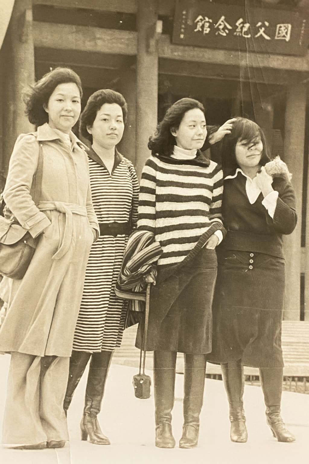A sepia photograph of four young women in 1970s coats and skirts, university friends, standing together in front of a memorial hall in Taipei.